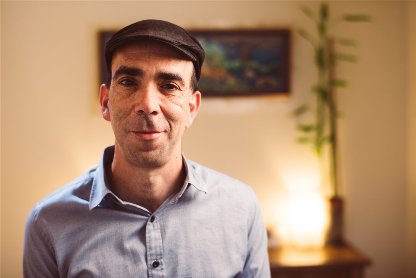 שאול זוארץ מחייך אל המצלמה, בכובע מצחייה וחולצה מכופתרת, בחדר מואר באור חם